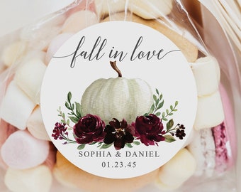 Puede incluir: Una bolsa transparente atada con una cinta blanca, llena de malvaviscos y galletas. Una pegatina redonda con el texto "fall in love" y un diseño de calabaza y flores en acuarela está adherida a la bolsa. Los nombres "SOPHIA & DANIEL" y una fecha también están impresos en la pegatina.