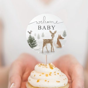 Op de afbeelding: Een witte cupcake met gouden sprinkles, bedekt met een ronde witte papieren cupcake topper met de tekst "welcome baby" met een hert, vos en vogels in een winters boslandschap.