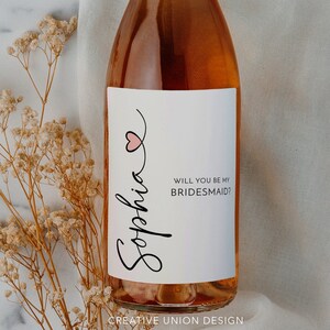 May include: A white wine bottle with a label that says "Sophia" in cursive with a pink heart and the text "Will you be my bridesmaid?" A bouquet of dried flowers is in the background.