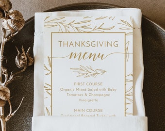 May include: A Thanksgiving menu card with gold floral accents is tucked into a white linen napkin on a dark gray plate. The menu features the words "Thanksgiving Menu" in elegant script, with course details below.