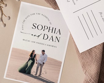 May include: Two white Save the Date postcards with black text. One card features a photo of a couple on a beach. The other card has the text "Save the Date" and contact information. The cards are on a tan cloth.