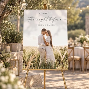 Könnte beinhalten: Ein rechteckiges Hochzeits-Willkommensschild auf einer goldenen Staffelei mit dem Text "WELCOME TO the night before SOPHIA & DANIEL" über einem Foto eines Paares in einem Feld mit hohem Gras. Das Schild hat einen weißen Hintergrund und schwarzen Text.