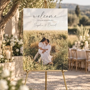 May include: A rectangular wedding welcome sign on a gold metal easel. The sign features a photograph of a couple sitting in a grassy field, with the text "welcome TO OUR BEGINNING" and the names "Sophia & Daniel" printed in black script.