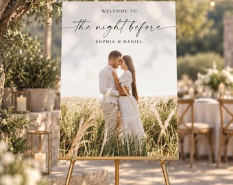 May include: A rectangular wedding welcome sign on a gold easel featuring the text "WELCOME TO the night before SOPHIA & DANIEL" above a photograph of a couple standing in a field of tall grass. The sign has a white background and black text.