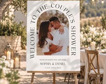 Puede incluir: Un letrero rectangular blanco para una fiesta de bodas sobre un caballete de madera con una foto arqueada de una pareja abrazándose. El texto dice WELCOME TO THE COUPLE'S SHOWER, WE'RE SO HAPPY YOU ARE HERE, SOPHIA & DANIEL, 01.23.45 • AUSTIN, TEXAS.