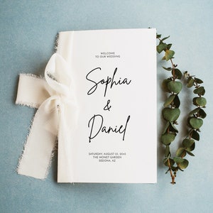 May include: A white wedding programme with the text "Welcome to our wedding" at the top, followed by the names "Sophia & Daniel" in cursive script. The date, location, and city are listed below the names. The programme is tied with a white ribbon and a sprig of greenery is to the right.