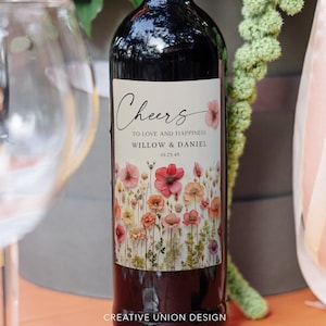 以下が含まれることがあります： 「Cheers to love and happiness Willow & Daniel 01.23.45」とパーソナライズされたラベルが付いた赤ワインのボトル。ラベルには、ピンク、白、黄色の花が咲く花柄のデザインが施されています。