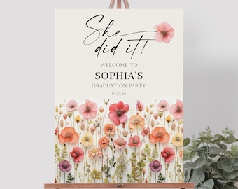 Puede incluir: Un cartel blanco con el texto "She did it!" en letra cursiva negra. Debajo del texto se lee "Welcome to Sophia's Graduation Party" en negro. El cartel está decorado con un borde floral de flores rosas, naranjas y amarillas.