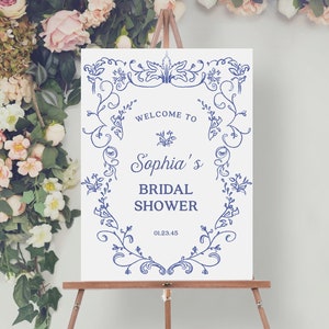 May include: White sign with blue floral border and text that reads "Welcome to Sophia's Bridal Shower 01.23.45". The sign is on a wooden easel.