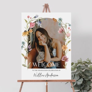 Puede incluir: Bienvenido a la celebración de graduación de Willow Anderson. Un cartel blanco con un borde floral y una foto de una persona sonriendo. El cartel está en un caballete de madera.