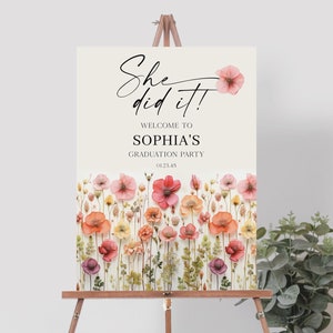 Puede incluir: Un cartel blanco con el texto "She did it!" en letra cursiva negra. Debajo del texto se lee "Welcome to Sophia's Graduation Party" en negro. El cartel está decorado con un borde floral de flores rosas, naranjas y amarillas.