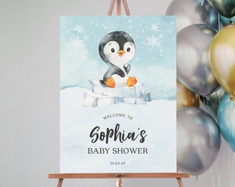 May include: A baby shower sign on a wooden easel features a watercolor illustration of a penguin on ice. The sign reads "Welcome to Sophia's Baby Shower" with the date 01.23.45. A cluster of balloons in silver, gold, and blue are in the background.