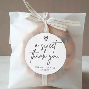 K&ouml;nnte beinhalten: Ein wei&szlig;er Papierbeutel mit einem silbernen Band, das um ihn herum gebunden ist. Der Beutel enth&auml;lt einen Keks mit einem runden wei&szlig;en Aufkleber, auf dem "a sweet thank you" mit einem Herz &uuml;ber dem Text steht. Der Aufkleber sagt auch "Sophia + Daniel 01.23.45"