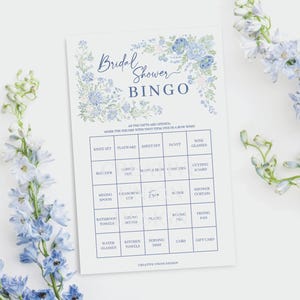 May include: A bridal shower bingo game card with a floral design in shades of blue. The card includes a grid with various gift-related items. The words "Bridal Shower Bingo" are at the top. The card is surrounded by blue flowers.