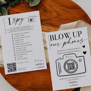 May include: Two white cards with black text on a wooden surface. One card says "I Spy" with a list of photo prompts and a QR code. The other says "Blow Up Our Phones" with a camera graphic and a QR code. Both cards say "Thank you, Sophia and Dan."