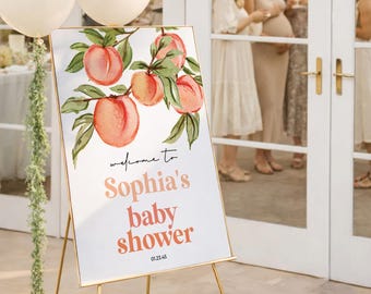 May include: A rectangular white sign with a gold frame on a gold tripod stand, featuring a watercolor peach illustration and the text: welcome to Sophia's baby shower 01.23.45. Three cream-colored balloons are attached to the side with green vines.