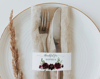 Puede incluir: Una mesa puesta con un plato blanco, una servilleta de lino beige y cubiertos. Una banda decorativa con el texto "thankful for SOPHIA" y detalles florales envuelve la servilleta. Un tallo de planta seca está a la izquierda.