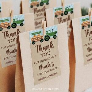 Op de afbeelding: Bruine papieren cadeauzakjes met labels. De labels tonen een groene tractor die een wagen met pompoenen trekt, met de tekst "Bedankt" en "Noah's Verjaardagsfeest". Houten knijpers bevestigen de labels aan de zakjes.
