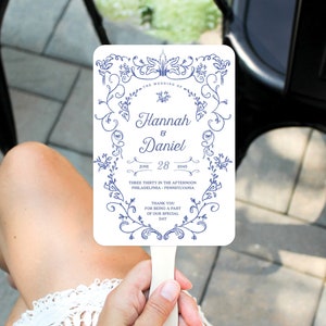 May include: A white wedding fan with a blue floral design. The fan features the text "The Wedding of Hannah & Daniel" with the date "June 28, 2045" and the time "Three Thirty in the Afternoon" along with the location "Philadelphia, Pennsylvania". The fan also includes the text "Thank You for being a part of our special day".