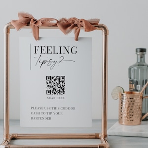 May include: A white sign with the text "FEELING Tipsy?" and a QR code. The sign is in a copper pipe frame with a brown ribbon. The text below the QR code reads "SCAN HERE PLEASE USE THIS CODE OR CASH TO TIP YOUR BARTENDER CREATIVE UNION DESIGN".