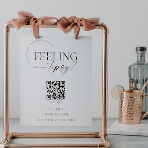 May include: A white sign with the text "Feeling Tipsy?" and a QR code with the text "Scan Here". Below the QR code is the text "Use this code or cash to tip your bartender". The sign is on a copper pipe stand with a brown ribbon.