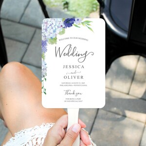 May include: A white wedding fan with a watercolor design of blue and purple hydrangeas and green leaves. The text on the fan reads "Welcome to our wedding, Jessica and Oliver, Saturday, August 23, 2045, Philadelphia, Pennsylvania, Thank you for being a part of our special day!"
