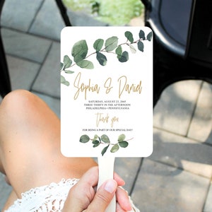 Może przedstawiać: Biały wachlarz ślubny z zielonymi akwarelowymi liśćmi eukaliptusa i złotym tekstem kaligraficznym. Tekst brzmi "Sophia & Daniel" i zawiera datę, godzinę i miejsce ślubu. Wachlarz mówi "Dziękuję" i "Za to, że byliście częścią naszego wyjątkowego dnia!"