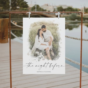 May include: A copper-colored metal stand holds a white sign with a watercolor-style image of a couple. The sign reads "the night before" and "SOPHIA & DANIEL". The background shows a lake and greenery.