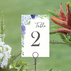 May include: A wedding table setting with floral arrangements, wine glasses, and a table number. The table number is white with blue floral accents and the number "2". A glass of red wine and a glass of white wine are visible.