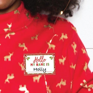 May include: A woman wearing a red long-sleeved shirt with a gold deer pattern, holding a champagne flute. The shirt has a high neck and a name tag that says "Hello, My Name is Holly." She is smiling, with dark curly hair and gold earrings.