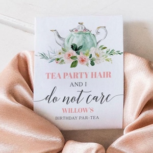 Może przedstawiać: Satynowa gumka do włosów w kolorze brzoskwiniowym z dołączoną białą kartką. Na kartce znajduje się akwarelowy rysunek czajnika z kwiatami i tekst "Tea Party Hair and I do not care Willow's Birthday Par-Tea."