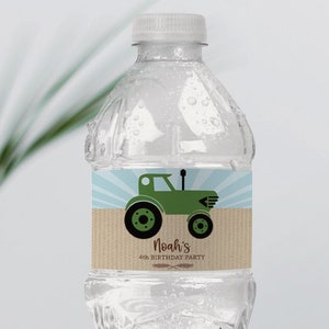 May include: A clear plastic water bottle with a white cap. The label features a green tractor illustration on a light blue and brown background. Text on the label reads "Noah's 4th Birthday Party". A clear vase with green leaves and a white camera are in the background.
