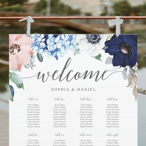 May include: A wedding seating chart with floral accents in pink, blue, and navy. The word "welcome" is written in cursive above the names "Sophia & Daniel." The chart is displayed on a copper-colored stand, with a lake in the background.