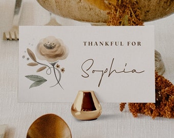 Puede incluir: Una mesa decorada con un centro de mesa floral en un cuenco metálico, una tarjeta con "THANKFUL FOR Sophia" y cubiertos dorados. Las flores son en tonos naranja, rojo y melocotón. También hay un pequeño candelabro dorado.