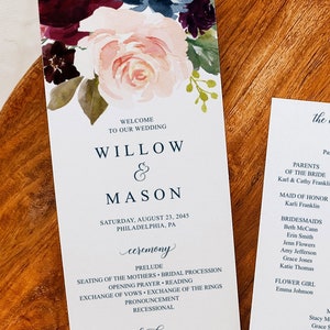 May include: Wedding ceremony programs with floral accents. The programs feature the names Willow & Mason, ceremony details, and a thank you message. The second program lists the wedding party and a honeymoon fund. The programs are on a wooden surface.