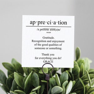 May include: A small potted plant with green leaves in a woven basket. A card with the word "appreciation" and its definition is displayed in a metal holder.