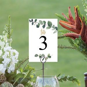 May include: Table number card with a white background and green eucalyptus leaves. The card has gold lettering that says "table 3".