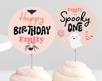 Op de afbeelding: Twee cupcakes met witte glazuur, bekroond met roze ronde decoraties. Eén zegt "Happy Birthday Emily" met vleermuis- en spinnengraphics. De andere zegt "Emily is a Spooky One" met spook- en snoepgraphics. Op een wit oppervlak.