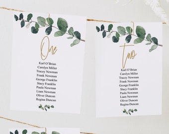 May include: Six white cards with green foliage and gold lettering, hanging from a string. Each card has a number from one to six and a list of names. The cards are for a wedding or event seating chart.