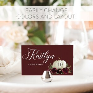 May include: A burgundy place card with the name "Kaitlyn" in elegant script, a white pumpkin illustration, and the number 10. The card is decorated with floral accents and sits on a white plate. The text "EASILY CHANGE COLORS AND LAYOUT!" is at the top.