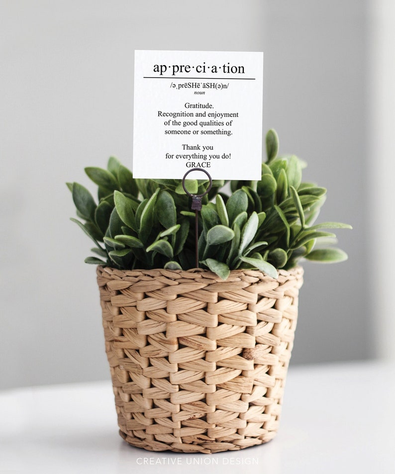 May include: A small potted plant with green leaves in a woven basket. A card with the word "appreciation" and its definition is displayed in a metal holder.