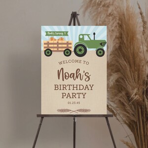 K&ouml;nnte beinhalten: Ein Geburtstagsparty-Schild auf einer Staffelei. Das Schild zeigt einen gr&uuml;nen Traktor, der einen Wagen mit K&uuml;rbissen zieht. Der Text lautet "Willkommen zu Noahs Geburtstagsparty" mit dem Datum 01.23.45. Das Schild ist mit Weizen&auml;hren verziert.