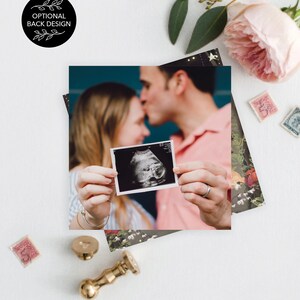 May include: A couple holds an ultrasound image of their baby in front of a teal background. The image is in a white frame and shows a baby in the womb. The couple is smiling and looking at the image. The image is surrounded by a floral pattern.