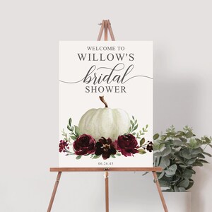 May include: Welcome to Willow's bridal shower sign with a white pumpkin and burgundy floral arrangement. The date 06.26.45 is printed below the floral arrangement.