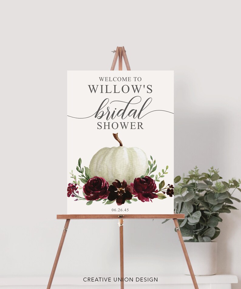 May include: Welcome to Willow's bridal shower sign with a white pumpkin and burgundy floral arrangement. The date 06.26.45 is printed below the floral arrangement.