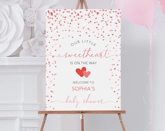 May include: A white baby shower sign with pink and red hearts and confetti. The sign says "Our Little Sweetheart is on the Way. Welcome to Sophia's Baby Shower."