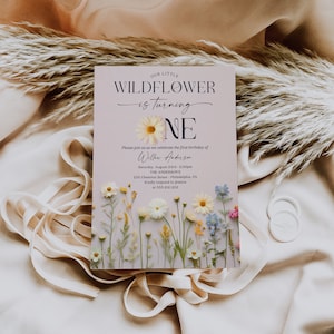 May include: A purple and white invitation card with a floral design. The text reads "Our Little Wildflower is turning One. Please join us as we celebrate the first birthday of Willow Anderson. Saturday, August 23rd, 2:00pm. The Anderson's 235 Chestnut Street, Philadelphia, PA. Kindly respond to Jessica at 555.212.1212. Creative Union Design."