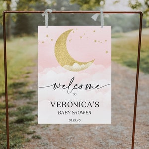 May include: A rectangular sign featuring a pink watercolor background with a gold glitter crescent moon, white clouds, and small gold stars. The text reads: welcome TO VERONICA'S BABY SHOWER 01.23.45. The sign hangs from a copper-colored metal frame.