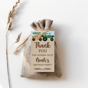 Op de afbeelding: Een kleine jute cadeauzak met een label met de tekst "Bedankt" voor Noah's verjaardagsfeestje. Het label heeft een groene tractorafbeelding en de tekst "Noah wordt 4". Het label is met touw aan de zak bevestigd.