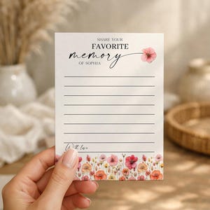 May include: A floral memory card with the text "Share Your Favorite Memory of Sophia." The card has a watercolor floral border with pink, orange, and yellow flowers. The card is held by a hand.